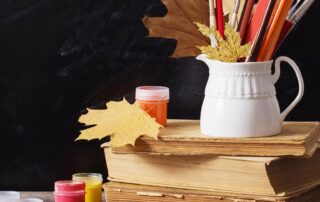 Schoolbooks on top of a desk with a penholder with pens, pencils and coloring pencils, small plastic paint pots, and a few autumn leaves