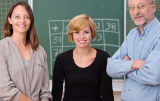 Group of three school teachers with confident friendly smiles standing in front of a class blackboard, one man and two women