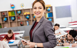 Smiling teacher, standing in front of the classroom, holding a digital table, with children writing notes in their desks